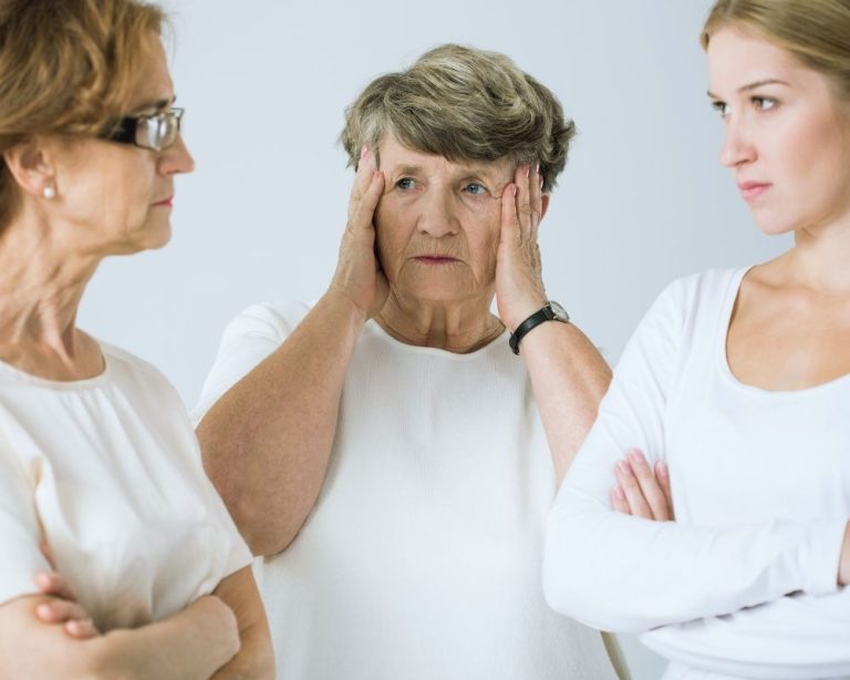 Zwei Frauen stehen sich mit verschränkten Armen gegenüber, im Hintergrund hält sich eine ältere Frau die Hände an den Kopf – Symbolbild für fremde Einflüsse auf unser Leben