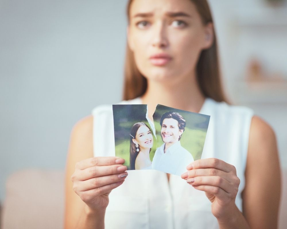 Eine Frau hält ein zerrissenes Foto eines Paares in den Händen – Symbolbild für eine negative Verbindung, die sie noch immer an eine vergangene Beziehung bindet